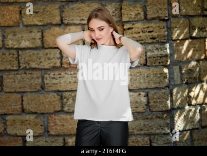 Vorlage eines weißen T-Shirts auf einem Mädchen, das ihre Haare korrigiert, auf dem Hintergrund einer Ziegelmauer mit Schatten, Vorderansicht. Produktfotografie. Modell von Stockfoto