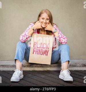 Die lächelnde blonde Frau sitzt auf dem Boden und hält eine Einkaufspapiertüte mit einem Black Friday Schild, während sie in die Kamera schaut Stockfoto