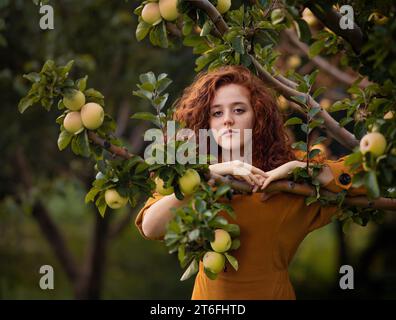 Porträt eines lockigen, schönen Mädchens in rotem Samtkleid und Schal, der im Apfelgarten posiert. Kunstwerk einer romantischen Frau. Hübsches Zärtlichkeit-Modell, das aussieht Stockfoto