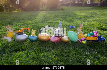 Metallmodelle von Spielzeugbaggern und kleinen Lkw stehen auf bunten Eiern unterschiedlicher Größe auf Gras. Geschäftskonzept für Ostergrüße für Konstrukt Stockfoto