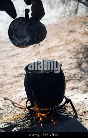 Ein Mann (Kamel-Trekking-Führer Nasser) gießt Wasser in einen großen Topf über einem Feuer in der Wüste. Stockfoto