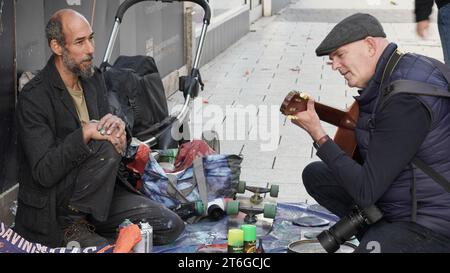 Ein Straßenkünstler, Finger in Sprühfarbe eingeklemmt, plaudert mit einem Straßenfotografen, der die Gitarre spielt, die er für Busking benutzt, und sich gegenseitig Gesellschaft genießt Stockfoto