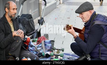 Ein Straßenkünstler, Finger in Sprühfarbe eingeklemmt, plaudert mit einem Straßenfotografen, der die Gitarre spielt, die er für Busking benutzt, und sich gegenseitig Gesellschaft genießt Stockfoto