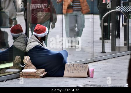 Elend im Stadtzentrum von Cardiff. Ein Obdachloser mit einem Schild und einem weihnachtsmann-Hut sitzt unter einer Decke in der Eingangspassage von John Lewis Betteln Stockfoto