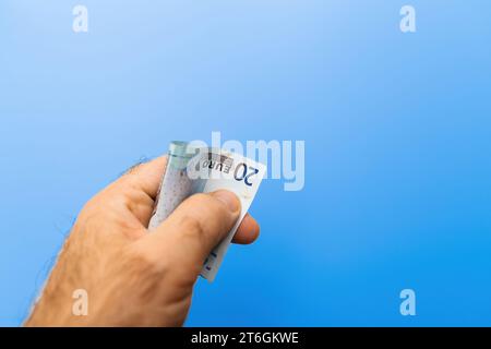 Ein Nahbild, das eine Hand mit einer gefalteten 20-Euro-Banknote zeigt. Die Hand wird auf einem durchgehend blauen Hintergrund angezeigt, wobei der Fokus auf der Währung liegt Stockfoto