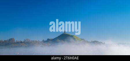 Ein Blick auf das Glastonbury Tor am frühen Wintermorgen vom Wearyall Hill mit Nebelbänken rund um das Tor und einem klaren blauen Himmel dahinter. Stockfoto