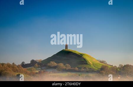 Ein Blick auf das Glastonbury Tor am frühen Wintermorgen vom Wearyall Hill mit Nebelbänken rund um das Tor und einem klaren blauen Himmel dahinter. Stockfoto