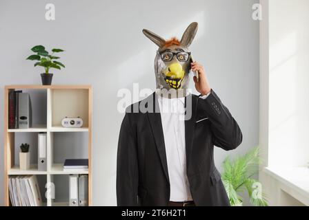 Lustiger Mann mit Tieresel-Maske auf dem Kopf, der im Büro steht und mit dem Handy spricht. Stockfoto