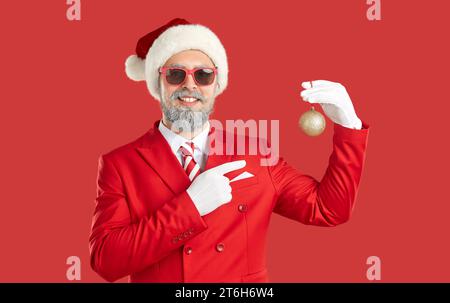 Mann in rotem Anzug, Hut und Sonnenbrille, der auf rotem Hintergrund steht und Weihnachtsball hält Stockfoto