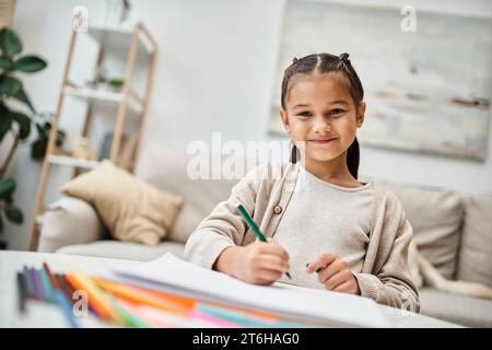 Porträt eines glücklichen Mädchens im Grundalter, das mit Farbstift zeichnet und in einem modernen Apartment lächelt Stockfoto
