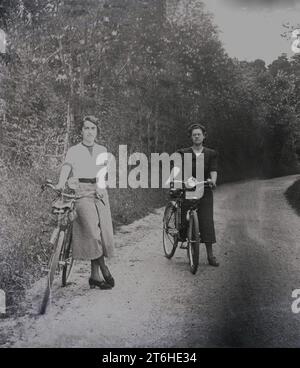 1950er Jahre, historisch, zwei junge Frauen in langen Röcken, die draußen auf einer ländlichen Gasse mit ihren Fahrrädern stehen, England, Großbritannien. Stockfoto