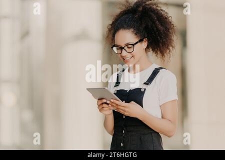 Verstrickte junge Frau mit lockigem Haar mit einer Tablette, lächelnd in einem stilvollen Outfit. Stockfoto