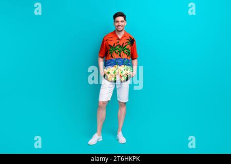 Foto in voller Größe des zufriedenen angenehmen Mannes tragen tropisches Hemd stilvolle Shorts Stehende Arme in den Taschen isoliert auf blauem Hintergrund Stockfoto