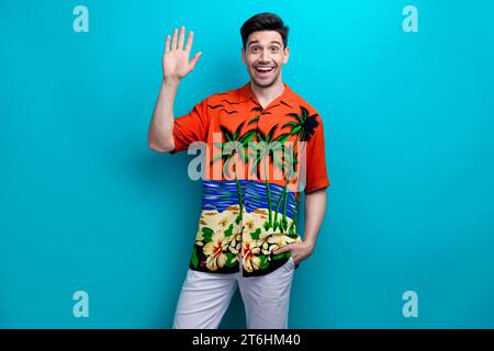 Foto des jungen Touristen Guy winkend Palme fünf Hallo Symbol tragen tropisches Druckhemd Sommer warme Feiertage isoliert auf blauem Farbhintergrund Stockfoto