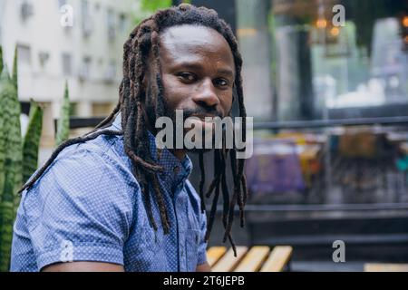 Profilansicht Porträt eines afrikanischen Mannes mit Bart, Dreadlocks und Hemd, der vor dem Café sitzt und die Kamera allein und ruhig betrachtet, Kopierraum. Stockfoto
