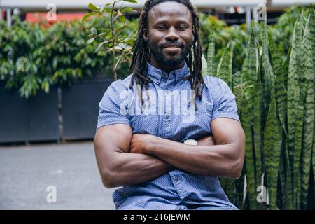 Junger Mann mit Dreadlocks und Bart in legeren Kleidern, der mit überkreuzten Armen vor dem Café sitzt, glücklich und lächelnd in die Kamera schaut Stockfoto