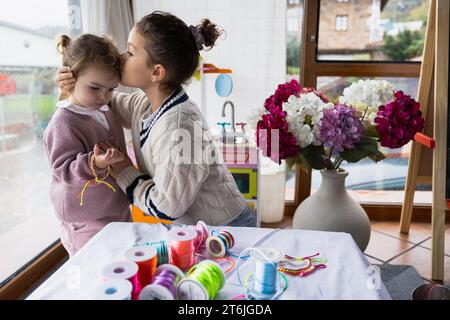 Zwei kleine Mädchen sitzen im Wohnzimmer und spielen mit bunten Fäden, die ältere küsst ihre Schwester Stockfoto