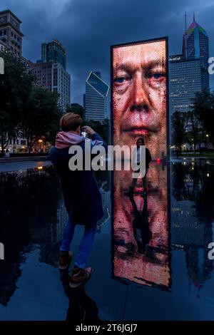 Ein Mann fotografiert eine Frau, die vor der Kunstinstallation „Gesichter von Chicago“ (Crown Fountain) im Millennium Park posiert, wobei das Gesicht eines digitalen Mannes auf seinem Turm leuchtet. Der katalanische Künstler Jaume Plensa schuf die 15,2 Meter (50 Fuß) hohen Glasziegel- und LED-Bildschirmtürme im Jahr 2004. Sie zeigen die wechselnden Gesichter von etwa 1.000 Chicagoern. Das Schild der Prudential-Versicherungsgesellschaft ist rechts im Hintergrund zu sehen. Chicago, Illinois, Vereinigte Staaten. Stockfoto