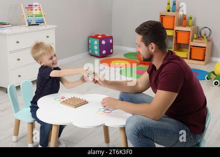 Motorische Entwicklung. Glücklicher Vater hilft seinem Sohn, mit Geoboard und Gummibändern an einem weißen Tisch im Zimmer zu spielen Stockfoto