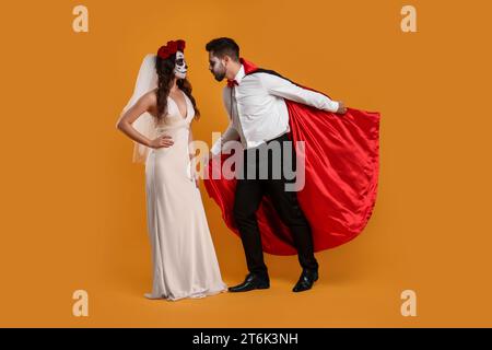 Ein Paar in gruseligen Braut- und Vampirkostümen auf orangem Hintergrund. Halloween-Feier Stockfoto