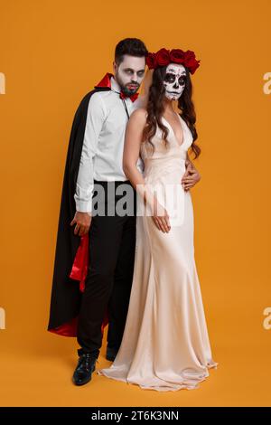 Ein Paar in gruseligen Braut- und Vampirkostümen auf orangem Hintergrund. Halloween-Feier Stockfoto