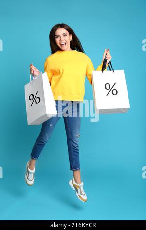 Rabatt, Verkauf, Angebot. Frau, die mit Einkaufstaschen vor hellblauem Hintergrund springt. Papiertüten mit Prozentzeichen Stockfoto