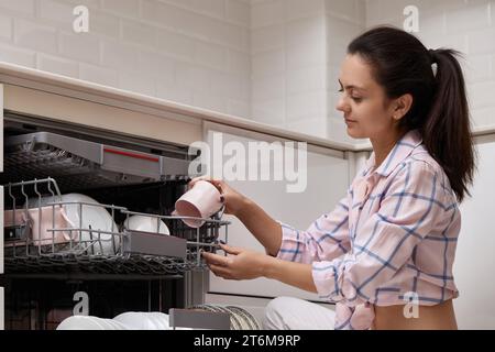 Attraktive Frau Entladebecher aus offener, eingebauter Spülmaschine mit sauberen Utensilien in der modernen weißen Küche. Stockfoto