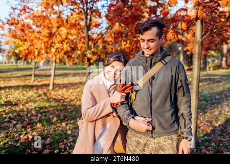 Junger Soldat in Uniform und seine Frau im Herbstpark. Das Paar hält nach langer Trennung die Hände. Lächelnde Frau lehnt sich auf die Schulter. Homecoming Stockfoto