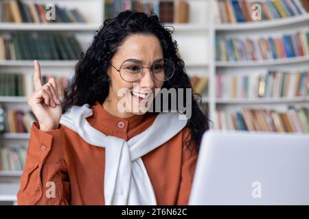 Ideenkonzept. Eine aufgeregte junge Indianerin mit Brille, die am Schreibtisch sitzt und mit einem Laptop ihren Zeigefinger in einem Klassenzimmer hochhebt. Staunte, schockierte Dame, sehend, aha Moment. Stockfoto