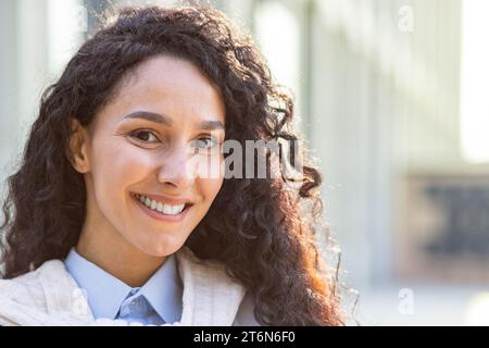 Nahaufnahme Porträt einer glücklichen schönen hispanischen Büroangestellten, die vor dem Büro bei Sonnenschein steht, einer erstaunlichen brünetten Studentin, die zufrieden in die Kamera blickt und lächelt. Stockfoto