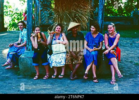 Indonesien. Bali. Touristen mit einheimischem Mann, der draußen unter einem Baum sitzt. Stockfoto