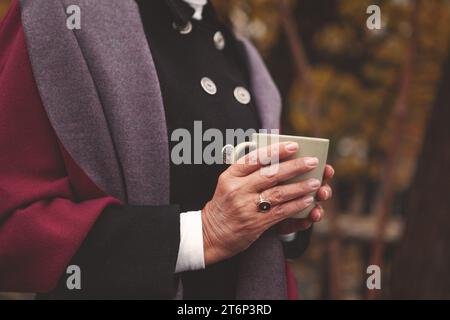Nahaufnahme der Hände einer älteren Frau, die bequem gekleidet ist und eine Tasse mit Kaffee oder Tee vor dem Hintergrund der herbstlichen Natur hält. Entspannende Atmosphäre, warme Atmosphäre Stockfoto