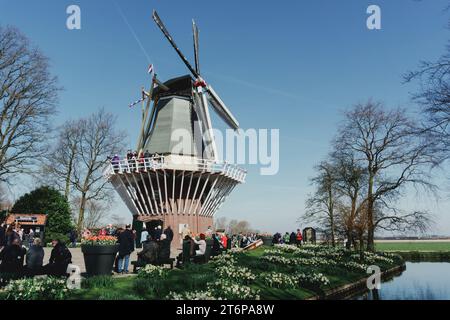 Eine traditionelle Windmühle steht hoch vor einem klaren Himmel, während eine Gruppe von Menschen einen angenehmen Tag am Ufer eines ruhigen Gewässers genießt Stockfoto