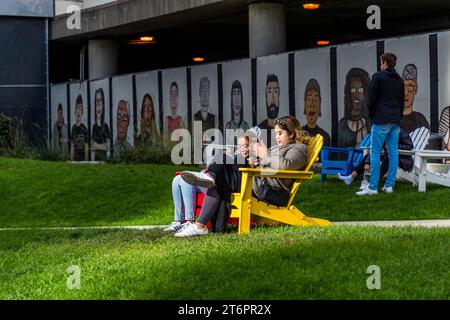 Chicago Riverwalk mit Grünflächen und Sitzgelegenheiten. Im Hintergrund Street Art von Straßenkünstler Don t Fret mit 55 Porträts von Chicagoern 'The People in your Neighborhood'. Die Menschen entspannen sich in Adirondack Stühlen auf dem Rasen des Chicagoer Riverwalk am Zusammenfluss der drei Zweige des Chicago River. Chicago, Usa Stockfoto