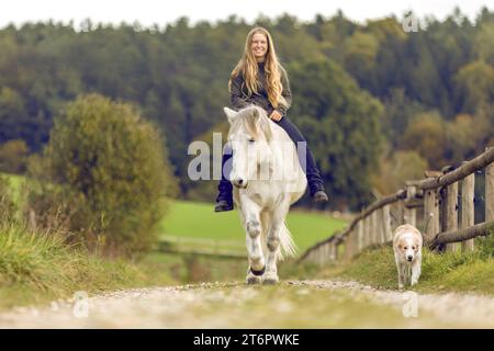 Pferd und Hund Konzept: Eine junge Reiterin und ihr Hund auf ihrem islandpferd vor ländlicher Bauernlandschaft Stockfoto