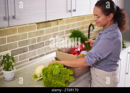 Charmante junge Frau, die einen gesunden Lebensstil pflegt, einen Karton mit frischen Bio-Lebensmitteln zu Hause auspackt, mitnimmt und sortiert Stockfoto