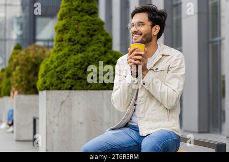 Glücklicher indischer Mann, der morgens Kaffee, heißes Getränk genießt und draußen lächelt. Entspannen, eine Pause einlegen. Ein Araber in der Innenstadt sitzt auf der Bank und trinkt Kaffee zum Mitnehmen. Stadtleben draußen Stockfoto