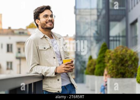 Glücklicher junger indischer Mann, der morgens Kaffee, heißes Getränk genießt und draußen lächelt. Entspannen, eine Pause einlegen. Ein arabischer Hindu-Typ in der Innenstadt, trinkt Kaffee zum Mitnehmen. Stadtleben draußen Stockfoto