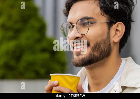 Nahaufnahme glücklicher indischer Mann, der morgens Kaffee, heißes Getränk genießt und draußen lächelt. Entspannen, eine Pause einlegen. Ein arabischer Hindu-Typ in der Innenstadt, trinkt Kaffee zum Mitnehmen. Stadtleben draußen Stockfoto