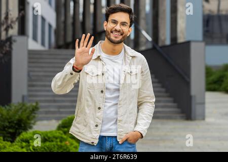 Hallo. Indischer Mann lächelt freundlich vor der Kamera, winkende Hände, die hallo, Begrüßung oder Abschied geben, einladend mit gastfreundlichem Ausdruck im Freien. Glücklicher arabischer Hindu-Typ, der in der Stadtstraße steht Stockfoto
