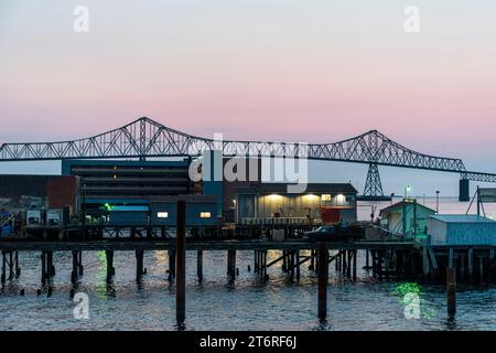 Ein funktionierender Pier und mehrere Gebäude nehmen den Vordergrund mit der Silhouette der Astoria-Megler Bridge vor einem rosafarbenen Nachthimmel im Hintergrund ein Stockfoto