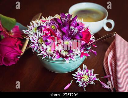 Rosafarbene und lila Blumen, die aus einer Frühstücksschale auf einem dunklen Tisch auslaufen. Tasse Kaffee im Hintergrund und Rose auf einem Beistellteller und einer gestreiften Serviette. Stockfoto