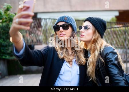 Zwei modische Frauen, die Mützen und Sonnenbrillen tragen, machen an einem sonnigen Tag ein Selfie, um einen Moment der Freundschaft festzuhalten. Stockfoto