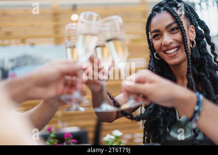Eine strahlende Frau mit geflochtenem Haar, die mit Sektgläsern unter Freunden bei einer Feier im Freien toast - Gruppe jubelt bei einem geselligen Treffen. Stockfoto