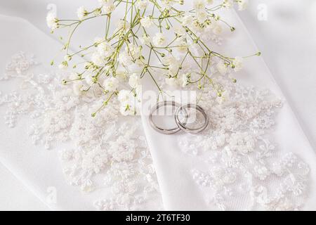 Zwei Eheringe aus Weißgold liegen auf den schicken Hochzeitshandschuhen der Braut. Weißer satinierter Hintergrund. Hochzeitszubehör-Konzept Stockfoto