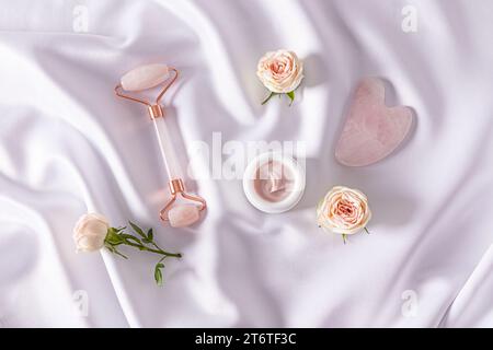 Set von Kosmetik und Werkzeugen auf weißem satiniertem Hintergrund mit lebenden Rosenknospen. Draufsicht. Kosmetische Massage. Häusliche Pflege. Stockfoto