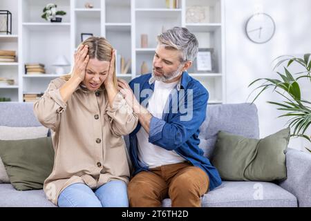 Seniorenpaar sitzt auf dem Sofa im Wohnzimmer, Frau reibt sich den Kopf mit den Händen, leidet an schweren Migräne-Kopfschmerzen, Ehemann umarmt sie, tröstet sie, bietet Hilfe an. Stockfoto