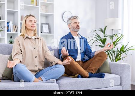 Seniorenpaar sitzt auf Sofa mit geschlossenen Augen in Lotusposition und übt Yoga im hellen Wohnzimmer. Sport zu Hause, ein gesunder Lebensstil, beruhigend und entspannend nach einem harten Arbeitstag. Stockfoto