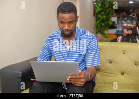 Fröhlicher afrikanischer Mann, der Computer benutzt und lächelt, während er auf dem Sofa sitzt. Konzept junger Geschäftsleute, die zu Hause arbeiten. Stockfoto