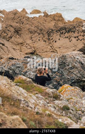 Eine Frau sitzt zwischen den Felsen und beobachtet Vögel durch ein Fernglas an der Küste in der Nähe von St Davids in Pembrokeshire in Südwales Stockfoto